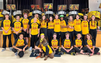 19 members of the band standing in front of their steel drums with a brown furry dog in a black and gold t-shirt